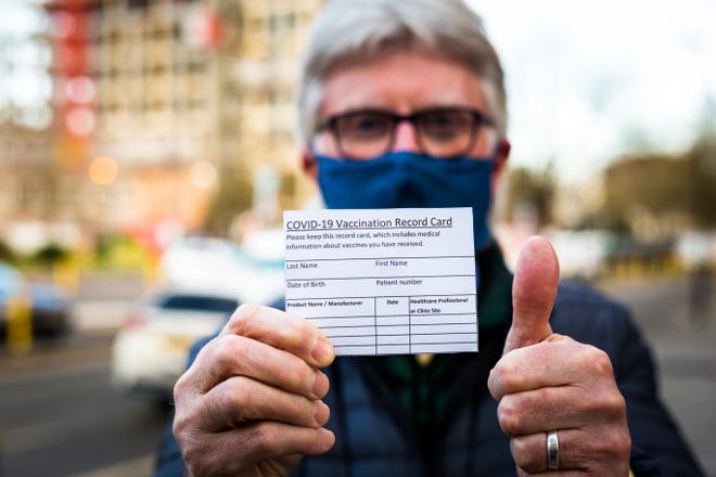 A man in a cloth mask, standing alongside a busy street, holding a COVID-19 vaccination card and giving a thumbs-up